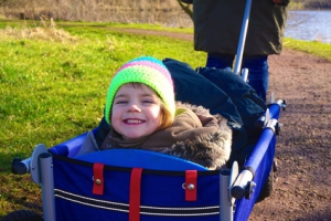 Kind sitzt lachend im Bollerwagen und lässt sich von der Mutter durch den Park ziehen.
